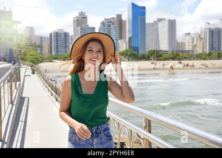 Ein stilvolles Mädchen mit Hut, das an einem windigen Tag entlang der Promenade spaziert, mit Wolkenkratzern im Hintergrund, Santos, Brasilien Stockfoto