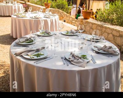 Wunderschöne Hochzeitsdekoration im Freien in der Stadt. Kerzen und getrocknete Blumen und Accessoires mit Blumensträußen und Gläsern auf dem Tisch mit Leinentischdecken auf dem Frischverheiratetstisch auf grünem Rasen Stockfoto