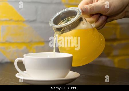 Gießt gelben Ingwertee mit Zitrone und Rosmarin-Zweig von Hand aus transparenter Teekanne in einen weißen Becher auf einem Holztisch Stockfoto