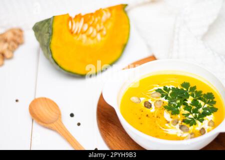 Ein Teller gelbe Kürbiscremesuppe mit Sahne, Kräutern und Samen auf einem weißen Tisch neben rohem Kürbis und einem Holzlöffel. Stockfoto
