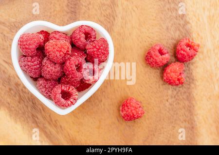 Herzförmige Schüssel voller reifer roter, saftiger Himbeeren auf Holzhintergrund. Draufsicht, Kopierbereich Stockfoto