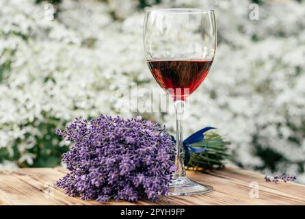 Ein Glas Rotwein und ein lavendelfarbener Blumenstrauß mit blauem Band auf einem Holztisch an sonnigen Tagen. Organischer Hintergrund. Weichzeichner Stockfoto