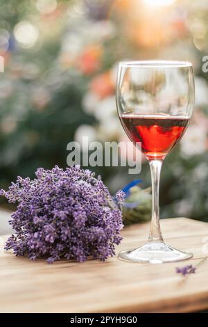 Ein Glas Rotwein und ein lavendelfarbener Blumenstrauß mit blauem Band auf einem Holztisch an sonnigen Tagen. Organischer Hintergrund. Weichzeichner Stockfoto