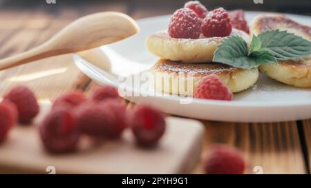 Köstliche Käsekuchen oder Pfannkuchen mit Puderzucker auf weißem Teller auf einem Holztisch mit Himbeeren und Minzeblättern Stockfoto