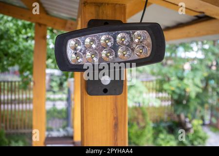 Nahaufnahme eines LED-Scheinwerfers, eines Scheinwerfers auf einem Holzpfosten einer offenen Veranda, auf einem unscharfen Hintergrund des Gartens. Eine Laterne zur Beleuchtung des Wohngebiets bei Nacht Stockfoto