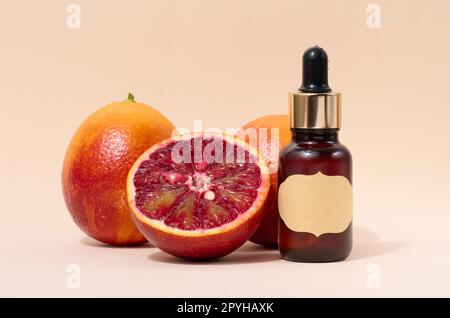 Braune Glasflasche mit einer Pipette und einem Papieretikett und ein halbes rotes Orange auf beigem Hintergrund, Öle und Kosmetika Stockfoto