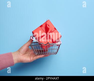 Die Hand einer Frau hält eine rote Geschenkbox, die in der Luft mit blauem Hintergrund verpackt ist, das Konzept der Feier, Überraschung Stockfoto