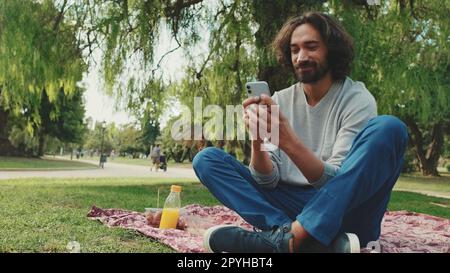 Junger Mann, der auf dem Rasen im Park auf dem Handy sitzt Stockfoto