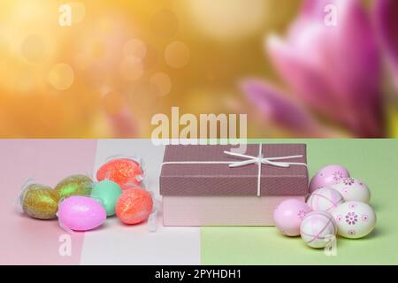 Vorlage für Ostergrußkarte. Nahaufnahme von bunten Eiern und einer Geschenkbox auf einem mehrfarbigen Tisch vor abstraktem Frühlingshintergrund. Kopierbereich für Ihr Textdesign. Stockfoto