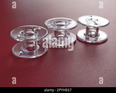 Drei transparente Nähmaschinenspulen aus Kunststoff. Textilindustrie. Detaildarstellung der Nähausrüstung. Roter Hintergrund, Hintergrundbeleuchtung Stockfoto