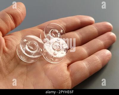 Transparente Kunststoffspulen einer modernen Nähmaschine in der Handfläche einer Frau - eine Näherin. Grauer Hintergrund. Nähmaschinen Stockfoto