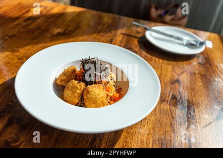 Fusionspaghetti garniert mit Ebiko und Seetang mit gebratenem paniertem Fleisch. Auf einem weißen runden Teller auf einem Holztisch schien Sonnenlicht durch das Fenster. Selektiver Fokus. Stockfoto