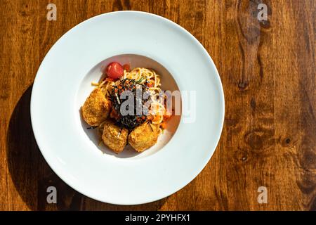 Draufsicht auf Fusionspaghetti, garniert mit Eiko und Seetang mit gebratenem paniertem Fleisch. Auf einem weißen runden Teller auf einem Holztisch schien Sonnenlicht durch das Fenster. Selektiver Fokus. Stockfoto