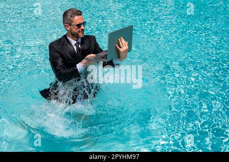 Ein lustiger, überraschter Geschäftsmann im Anzug mit einem Laptop im Swimmingpool. Verrückter, aufgeregter Geschäftsmann im Sommerurlaub. Aufgeregter Geschäftsmann in Neoprenanzug aufgeregt Stockfoto