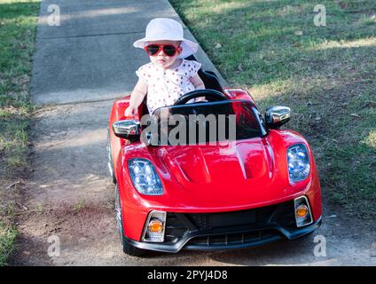 Ein weißes Baby mit weißem Hut, das in einem elektrischen Cabrio in rotem Sportwagen fährt. Ein Kind, das an einem Sommertag Auto fährt Stockfoto