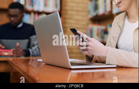 Vielleicht hilft mir eine Lerngruppe. Eine Universitätsstudentin, die ihr Handy benutzt, während sie hinter einem Laptop sitzt. Stockfoto