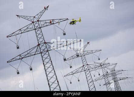 PRODUKTION - 03. Mai 2023, Schleswig-Holstein, Klixbüll: Ein Hubschrauber fliegt zwischen den Strommasten des letzten Bauabschnitts der Westküsten-Pipeline, während die so genannten Führungskabel eingezogen werden. Der Hubschrauber wird verwendet, um leichte Kunststoffkabel von einem Pol zum anderen zu fliegen, während industrielle Monteure das Führungskabel durch die Rollen an den Enden der Pole führen. Die Westküstenstrecke - eine 380-Kilovolt-Überlandleitung - wird über eine Gesamtlänge von etwa 140 km zwischen Brunsbüttel und der dänischen Grenze und von dort aus weiter verlaufen. Foto: Christian Charisius/dpa Stockfoto