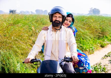 Glückliches junges indianisches Paar mit Schutzhelm auf Motorrad auf dem landwirtschaftlich genutzten Feld des Dorfes. Vorderansicht. Stockfoto