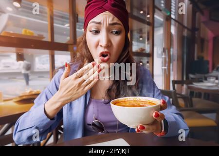 Eine indische Frau probiert eine scharfe und heiße Tom Yam Suppe in einem thailändischen Restaurant und reagiert emotional komisch. Gewürze und eine ungesunde Ernährung mit Over Stockfoto