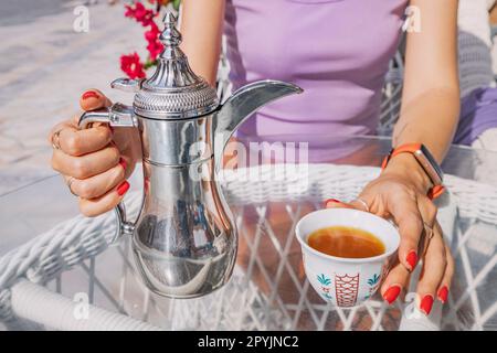 Mit den belebten Straßen von Dubai draußen, nimmt sich ein Touristenmädchen einen Moment Zeit, um den kräftigen Geschmack und das Aroma von frisch gebrühtem arabischem Kaffee von einem tra zu genießen Stockfoto