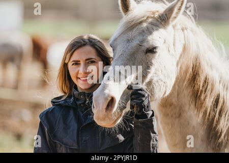 Junge Frau in schwarzer Reitjacke, die neben einem weißen arabischen Pferd steht und glücklich lächelt, Nahaufnahme Stockfoto