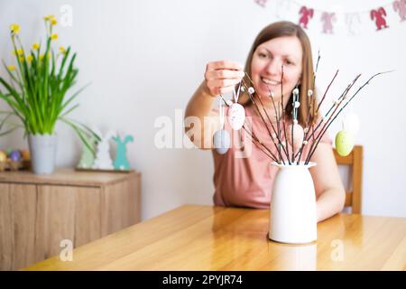 Lächelnde Frau, die zu Hause einen Strauß Weidenbrunches mit farbigen Ostereiern dekoriert. Stockfoto