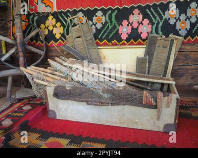 Antike Teppichwebwerkzeuge. Teppichflechten in Serbien und Bosnien. Handwerkskiste mit Holzrahmen, Drehrad. Holzhauswand mit farbenfrohen Teppichen. Antikes Bauernmotiv und Hobby Stockfoto