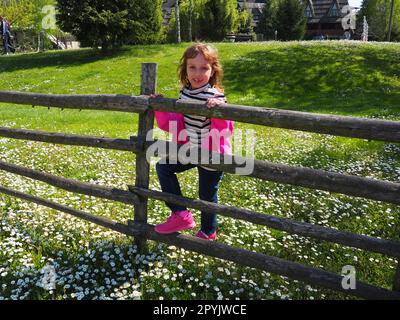 Stanisici, Bijelina, Bosnien und Herzegowina, 25. April 2021. Ein 7-jähriges Mädchen in einem rosa Pullover, Jeans und einer gestreiften Bluse steht neben einem hölzernen Zaun und lächelt. Wiese mit Wildblumen Stockfoto