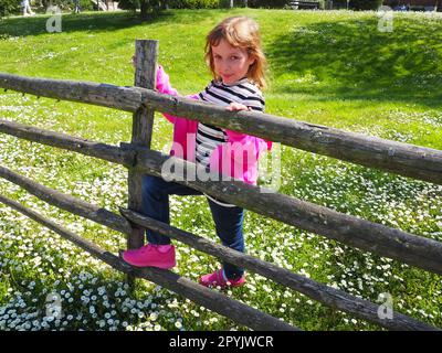 Stanisici, Bijelina, Bosnien und Herzegowina, 25. April 2021. Ein 7-jähriges Mädchen in einem rosa Pullover, Jeans und einer gestreiften Bluse steht neben einem hölzernen Zaun und lächelt. Wiese mit Wildblumen Stockfoto