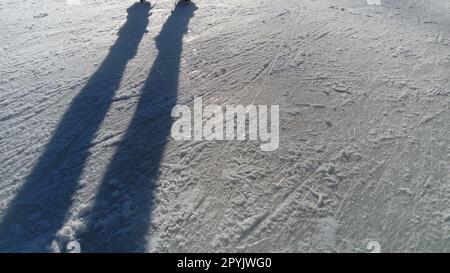 Kinder fahren in einem Stadtpark auf einer Eisbahn. Beim Eislaufen laufen laufen laufen die Füße. Die niedrige Wintersonne erhellt das Eis schwach. Dunkle Formen und lange Schatten auf der Oberfläche. Sportbewegungen Stockfoto