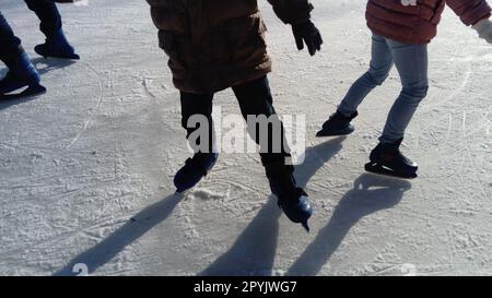 Kinder fahren in einem Stadtpark auf einer Eisbahn. Beim Eislaufen laufen laufen laufen die Füße. Die niedrige Wintersonne erhellt das Eis schwach. Dunkle Formen und lange Schatten auf der Oberfläche. Sportbewegungen Stockfoto