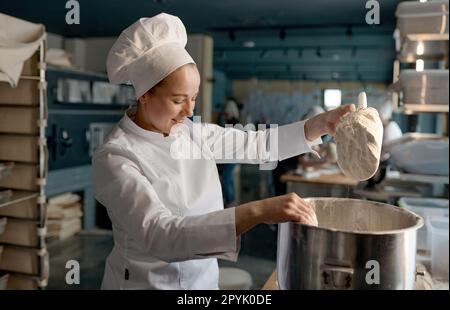 Weibliche Süßwarenfrau in weißer Uniform, die Mehl in eine große Metallschüssel schüttet Stockfoto