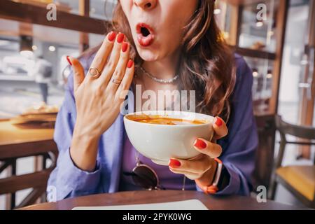 Das Mädchen probiert eine scharfe und heiße Tom-Yam-Suppe in einem Restaurant und reagiert emotional komisch. Gewürze in der nationalen Küche und eine ungesunde Ernährung mit ov Stockfoto