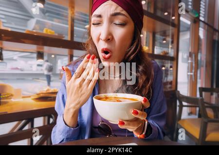 Eine indische Frau probiert eine scharfe und heiße Tom Yam Suppe in einem thailändischen Restaurant und reagiert emotional komisch. Gewürze und eine ungesunde Ernährung mit Over Stockfoto