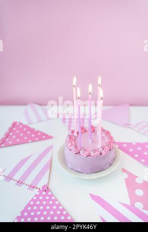 Zarter lavendelrosa Bento-Kuchen auf einem Teller mit pinkem Hintergrund mit Partyflaggen, Kerzen brennen auf dem Kuchen. Feier, fröhlicher Tag. Stockfoto