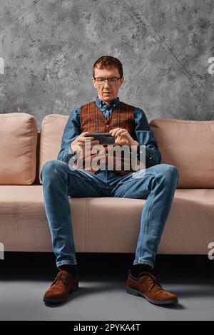 Ein Mann mittleren Alters, der auf dem Sofa sitzt und Videos auf dem Smartphone ansieht Stockfoto