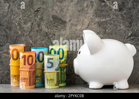 Weißes Keramik-Sparschwein, das auf die gerollten Euro-Banknoten schaut Stockfoto