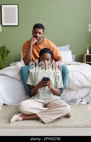 Afroamerikanisches junges Paar, das während der Freizeit im Schlafzimmer zu Hause ein Smartphone benutzt Stockfoto