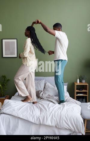Vertikales Bild eines jungen afroamerikanischen Paares, das in seiner Freizeit im Bett im Schlafzimmer tanzt Stockfoto