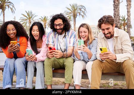 Fröhliche multikulturelle Gruppe von Freunden, die draußen auf einer Bank hintereinander sitzen und Mobiltelefone benutzen. Stockfoto