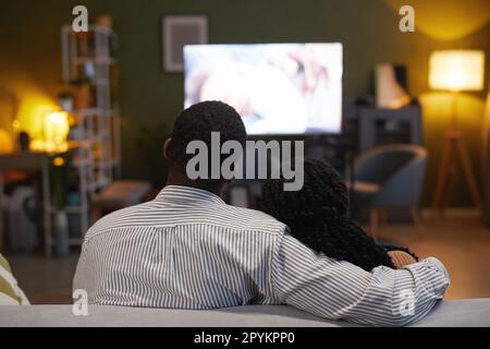 Rückansicht eines afroamerikanischen Paares, das sich umarmt, während es auf dem Sofa sitzt und zu Hause einen Film ansieht Stockfoto