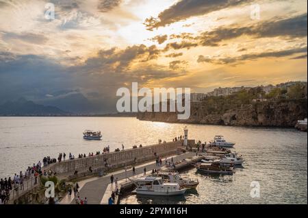 Menschen, die in der Dämmerung auf der Hafenmauer in Antalya, Provinz Antalya, Türkei (Turkiye) sitzen und laufen, während ein Vergnügungsboot zurückkehrt und der Himmel sich dreht oder Stockfoto