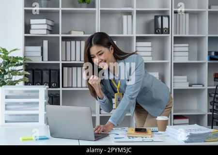 Eine junge Geschäftsfrau, die mit einem Notebook überglücklich war, die Freude im Büro zum Ausdruck brachte, und die aufgeregte Geschäftsfrau fühlt sich euphorisch, wenn sie im Internet gute Nachrichten liest Stockfoto