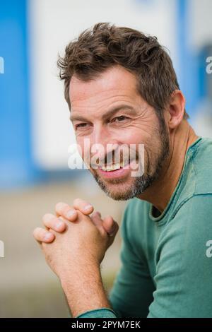 Porträt eines erwachsenen Mannes, der auf einer Bank vor der Backsteinwand eines weißen Gebäudes sitzt. Ein Mann, der sich ernsthaft ausgibt, sitzt auf einer Bank im Stree Stockfoto
