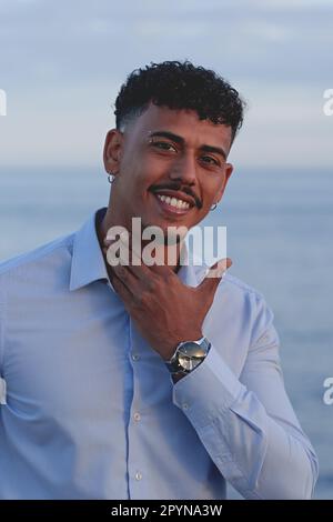 Porträt eines jungen afroamerikanischen Mannes mit lockigem Haar und Schnurrbart, lächelnd und berührend sein Kinn, mit Hemd und Uhr, mit dem Meer im Meer Stockfoto
