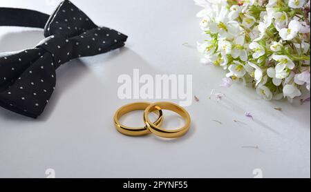 Hochzeitskonzept mit goldenen Ringen in der Mitte auf weißem Tisch und schwarzer Fliege und Blumenstrauß. Erhöhte Aussicht. Stockfoto