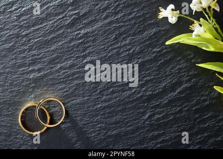 Hintergrund aus Schieferstein für die Hochzeitsankündigung mit zwei goldenen Ringen in einer Ecke und Blumen in einer anderen. Draufsicht. Stockfoto