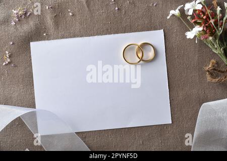 Hintergrund der Hochzeitsankündigung mit goldenen Ringen in Papierausschnitt auf braunem Tuch und Details mit weißem Band und Blumen. Draufsicht. Stockfoto