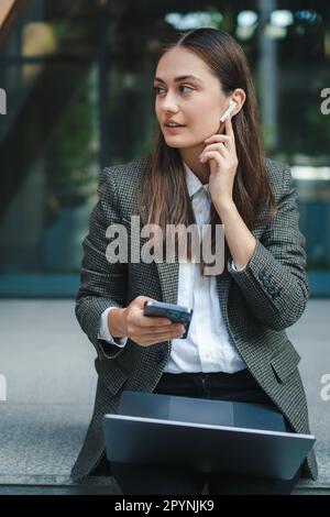 Attraktive junge brünette Frau, die mit ihrem Laptop auf der Treppe draußen auf dem Handy sitzt. Technologie, Kommunikation und Behaglichkeit Stockfoto