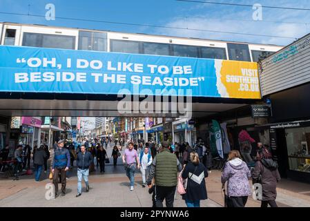Leute in der High Street, Southend auf dem Meer, die unter der Eisenbahnbrücke vorbeifahren mit "Oh, ich mag es, am Meer zu sein". Zug um C2C Uhr auf der Brücke. Beschäftigt Stockfoto